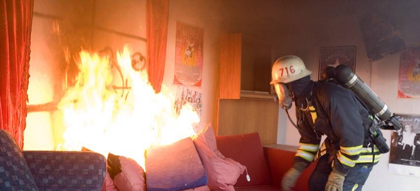 Syntolkning av bild: Brandman släckar brand i soffa