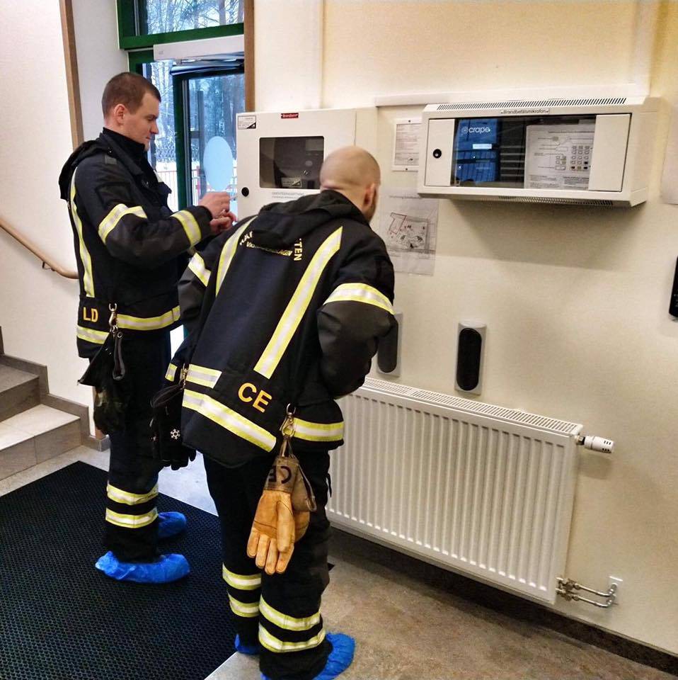 Syntolkning av bild: Brandmän som tittar på en tablå tillhörande en automatlarmsanläggning